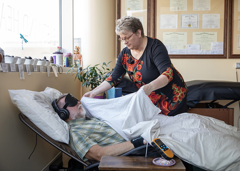 Dr. Husman tucks a blanket over a middle-aged man resting in a reclining zero gravity chair the man wears a sleep mask and headphones and medical monitor as he is prepared for a clinical ketamine or Spravato session.